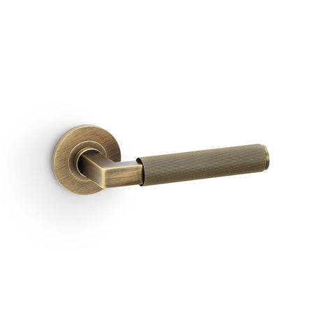 This is an image showing Alexander & Wilks Hurricane Reeded Lever on Round Rose - Antique Brass aw202ab available to order from Trade Door Handles in Kendal, quick delivery and discounted prices.