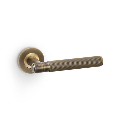This is an image showing Alexander & Wilks Spitfire Reeded Lever on Round Rose - Antique Brass aw222ab available to order from Trade Door Handles in Kendal, quick delivery and discounted prices.
