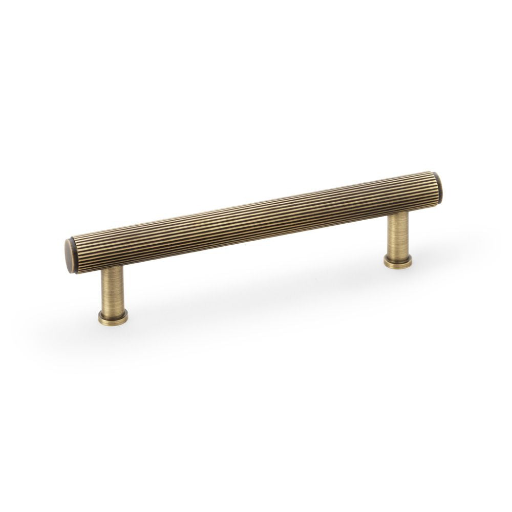 This is an image showing Alexander & Wilks Crispin Reeded T-bar Cupboard Pull Handle - Antique Brass - 128mm aw809r-128-ab available to order from Trade Door Handles in Kendal, quick delivery and discounted prices.