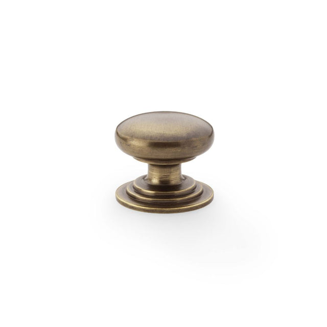 This is an image showing Alexander & Wilks Waltz Round Cupboard Knob on Stepped Rose - Antique Brass - 32mm aw825-32-ab available to order from Trade Door Handles in Kendal, quick delivery and discounted prices.