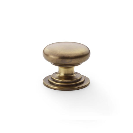 This is an image showing Alexander & Wilks Waltz Round Cupboard Knob on Stepped Rose - Antique Brass - 38mm aw825-38-ab available to order from Trade Door Handles in Kendal, quick delivery and discounted prices.