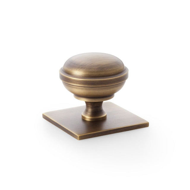 This is an image showing Alexander & Wilks Quantock Cupboard Knob on Square Backplate - Antique Brass - 38mm aw826-38-ab available to order from Trade Door Handles in Kendal, quick delivery and discounted prices.